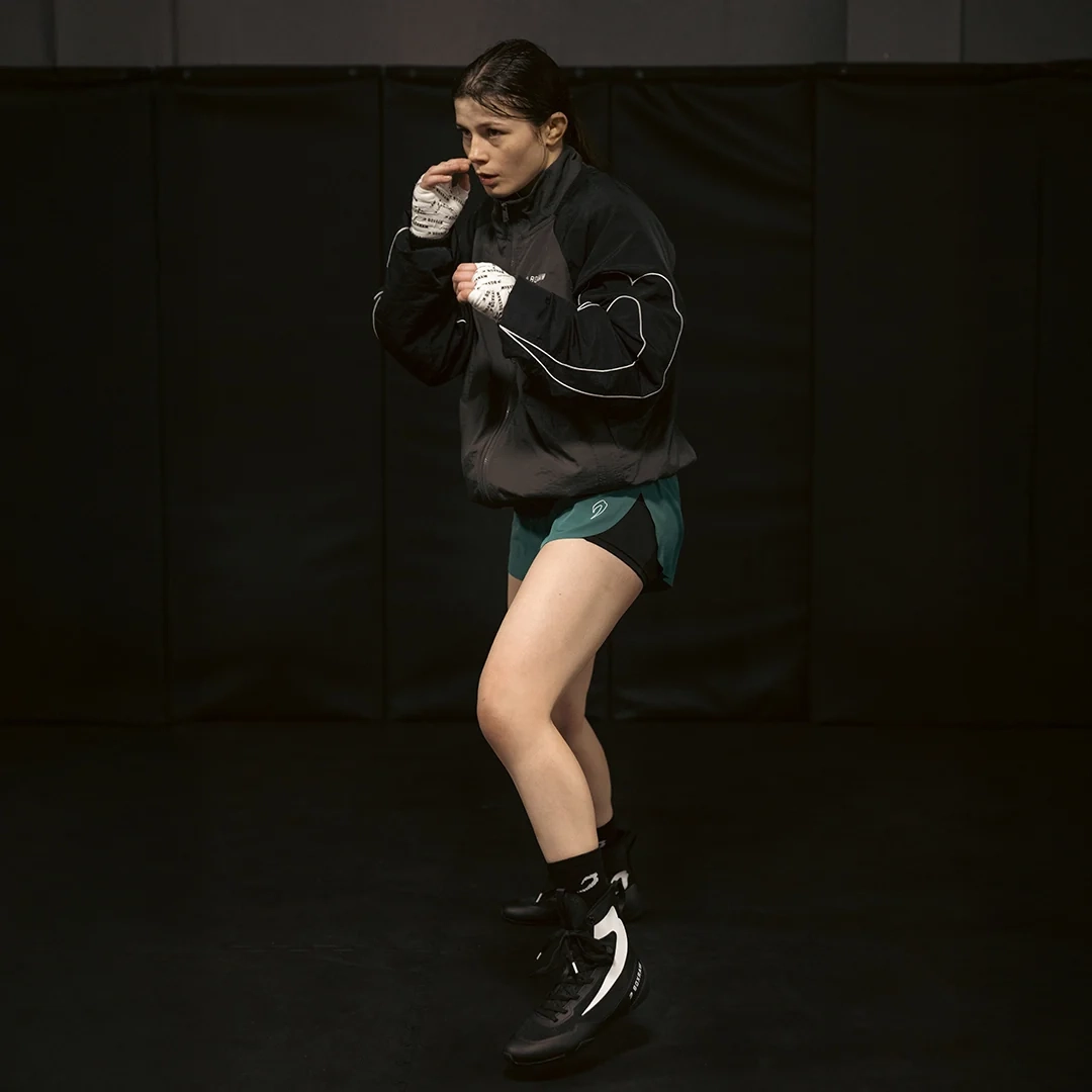 A woman in a black top and shorts shadowboxes against a dark wall