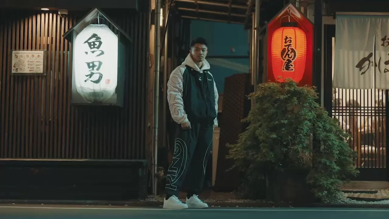 Two men in track suits stand in front of a lantern-lit doorway