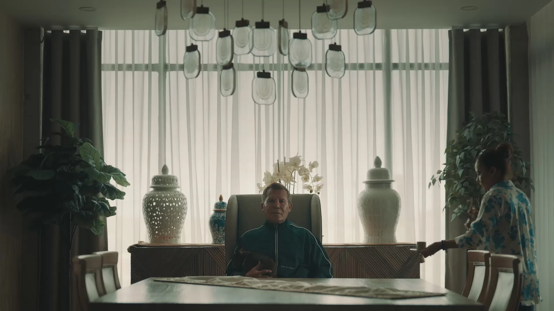 An older man in a dark track jacket sits at a long table in front of pale curtains