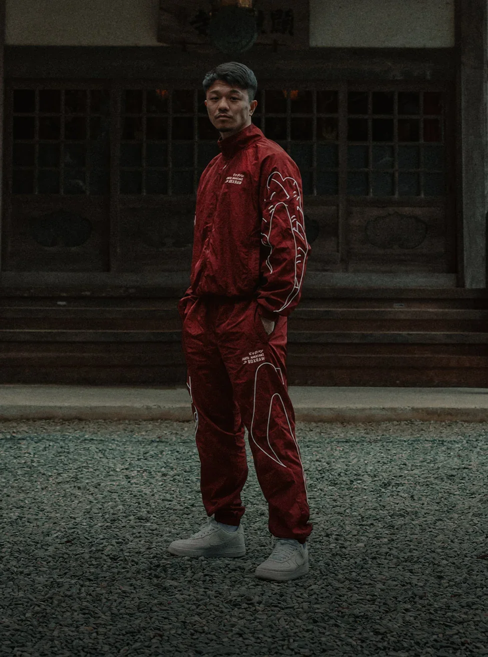 Nakatani standing in a red track suit in front of a dark wooden building