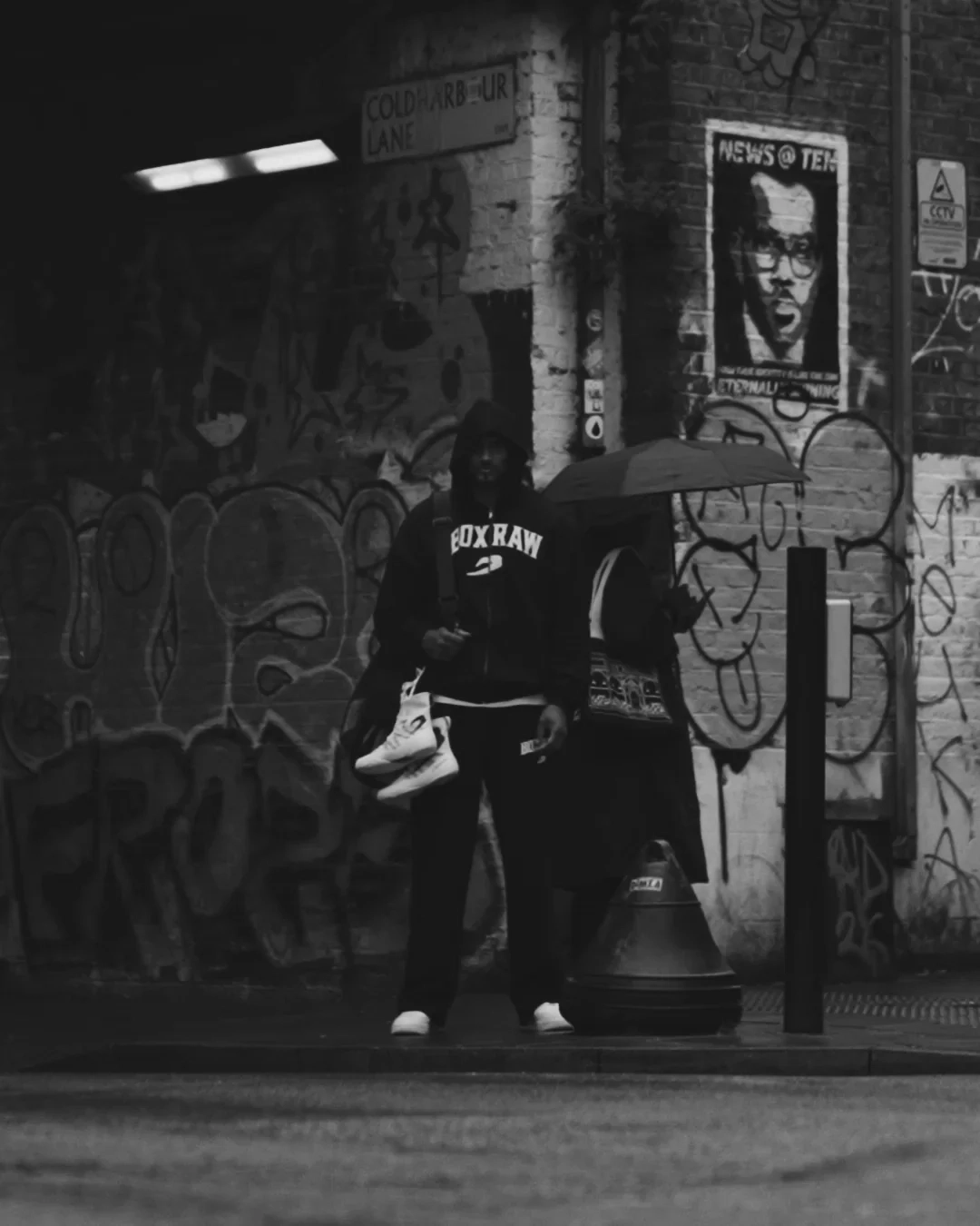 BOXRAW campaign still: a black-and-white alley scene with a hooded figure