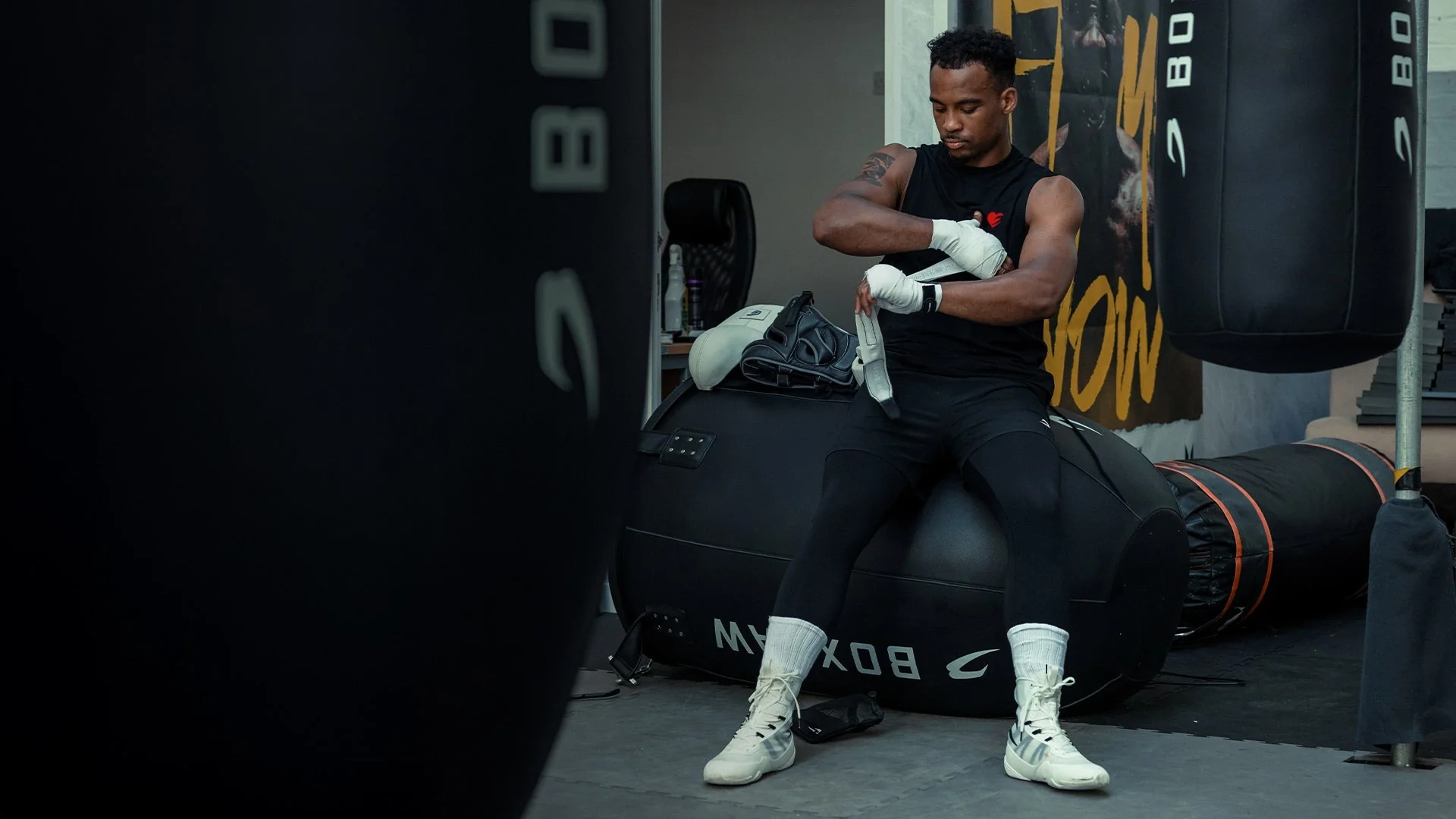 A boxer sits against black BOXRAW punch bags in a dark gym and wraps his hands